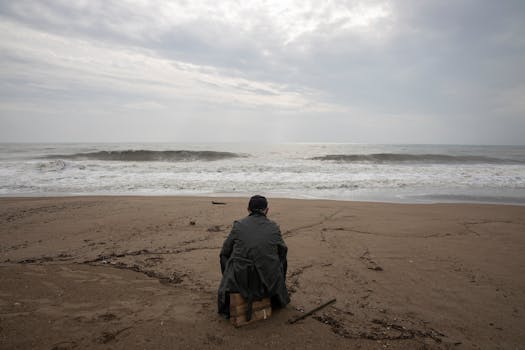 A solitary figure sits on a stormy beach, gazing at the turbulent sea under cloudy skies.